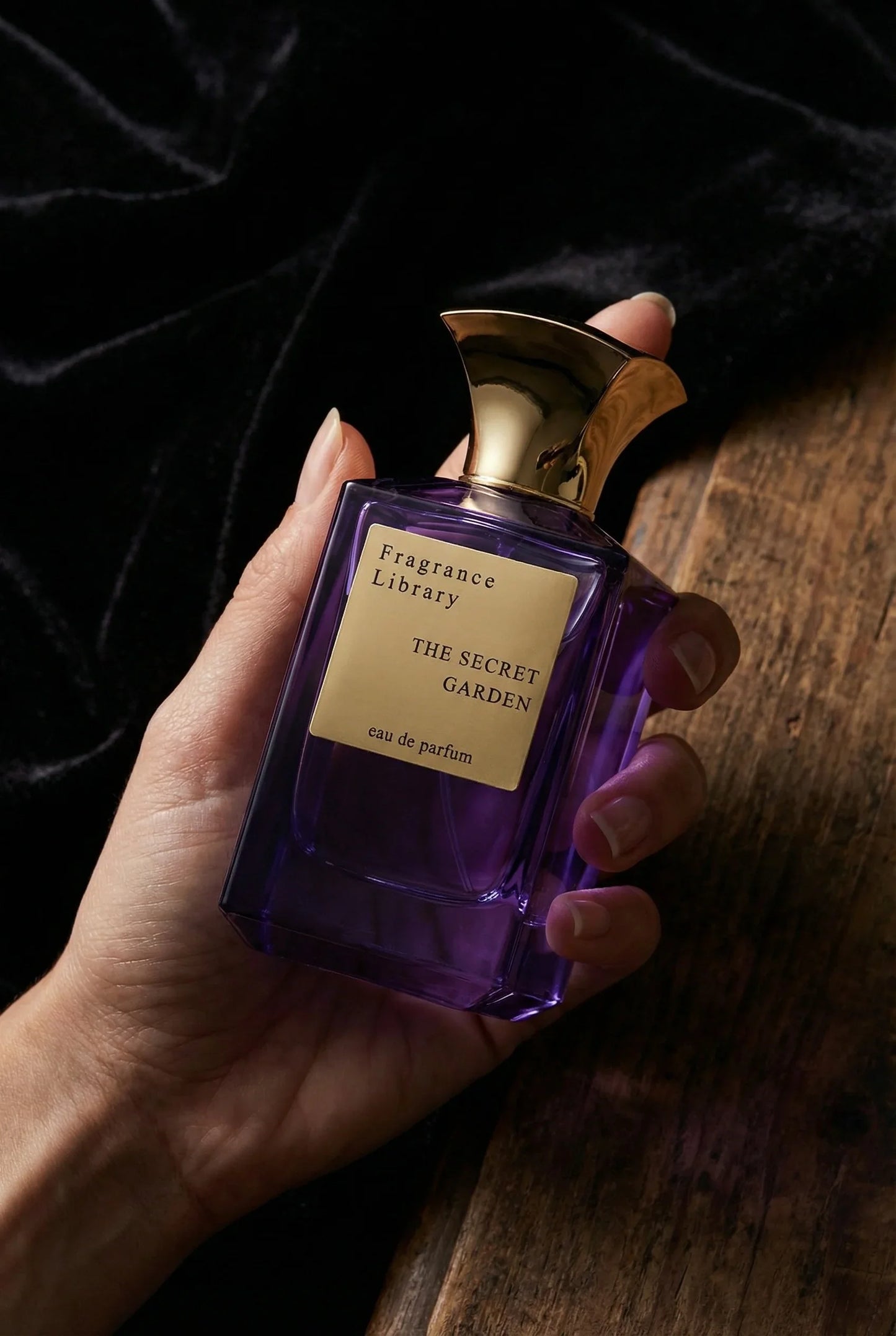 Lila Parfümflasche mit goldener Kappe und Label 'THE SECRET GARDEN' in Hand auf Holz