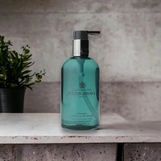 Molton Brown Coastal Cypress & Sea Fennel Handseife in türkisfarbener Flasche auf Marmoroberfläche, daneben grüne Pflanze