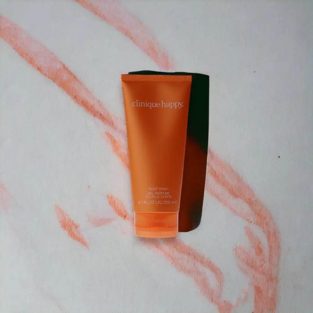 Orangefarbene Clinique Happy Duschgel Tube auf hellem Hintergrund mit rosa Linienmuster.