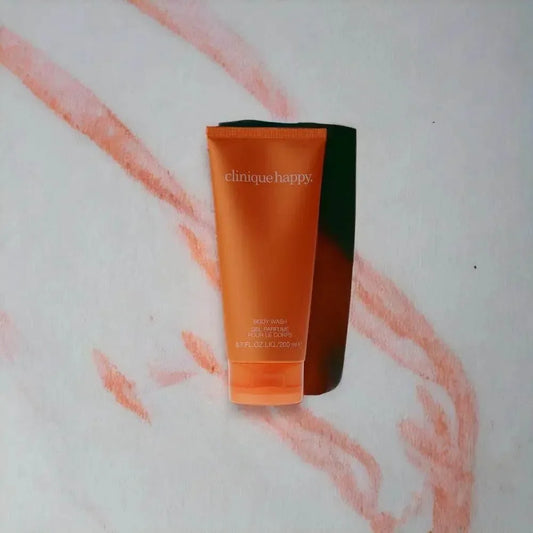 Orangefarbene Clinique Happy Duschgel Tube auf hellem Hintergrund mit rosa Linienmuster.