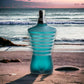 Türkise Parfumflasche in Torsoform mit Streifen, am Strand vor Meereskulisse
