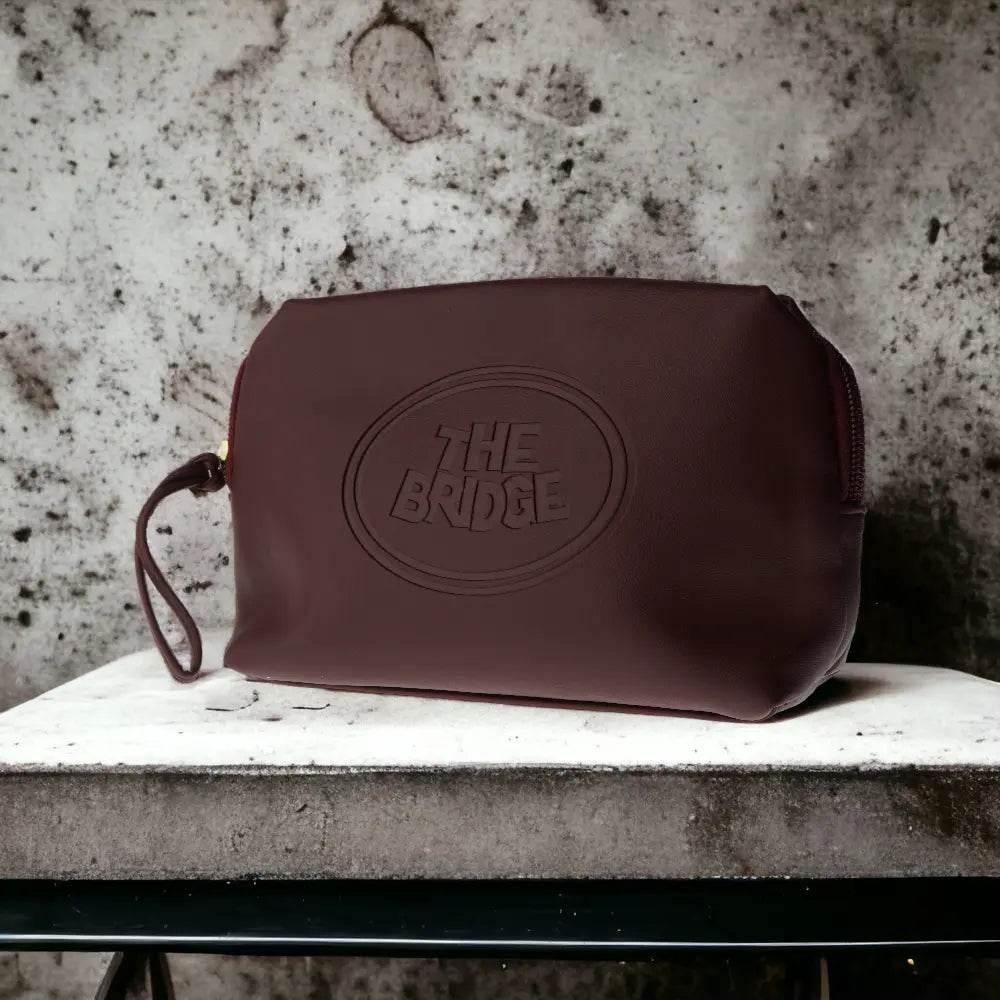 Bordeauxrote Kosmetiktasche aus Leder mit 'THE BRIDGE' Prägung, auf steinernem Tisch.
