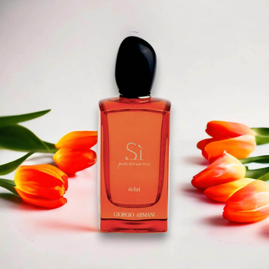 Rotes Parfumflakon Si Passione Éclat mit schwarzen Verschluss, umgeben von orange-gelben Tulpen, vor weißem Hintergrund.