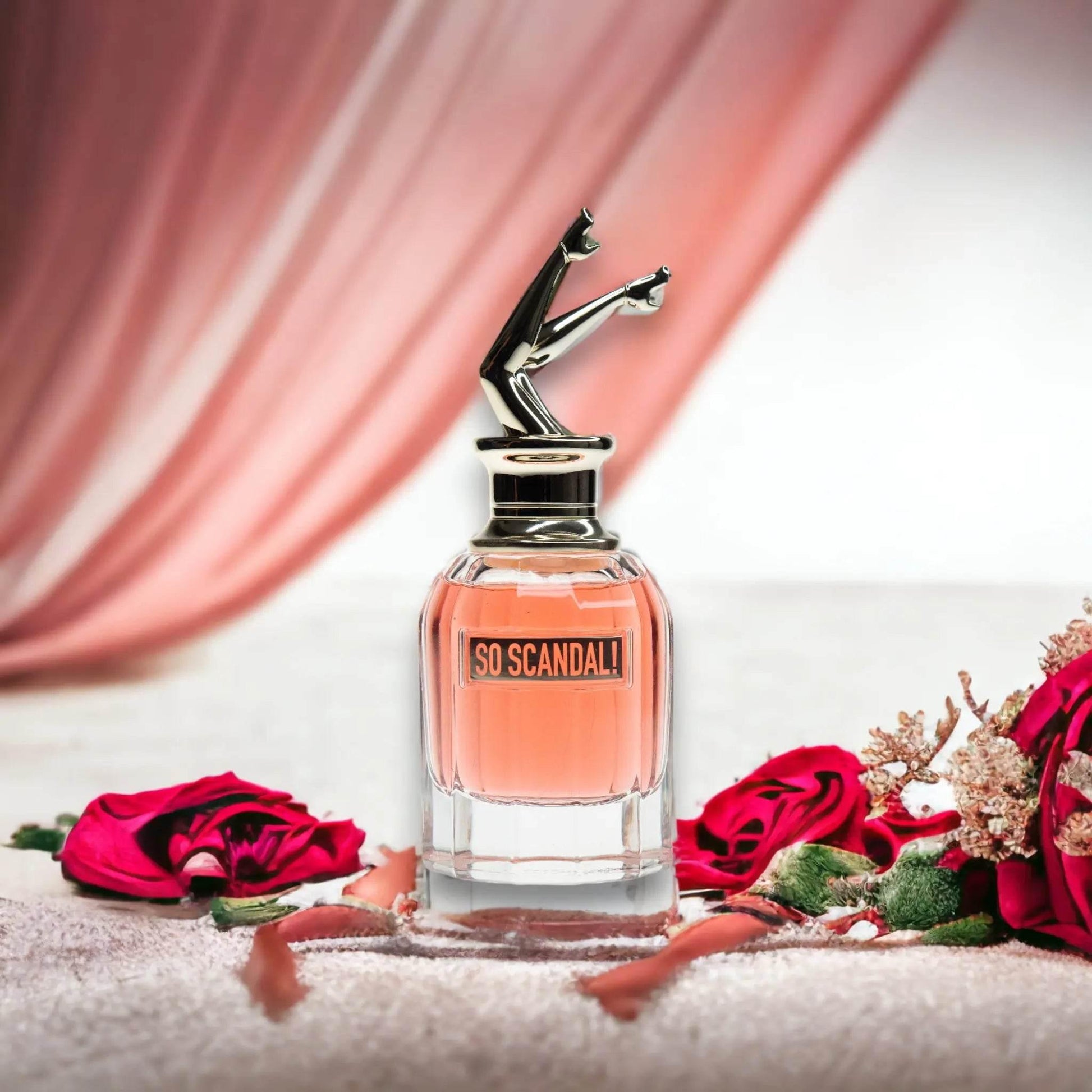 So Scandal Eau de Parfum Flakon mit rosa Duft, umgeben von Rosen und Schleifen, vor rosa Vorhang.
