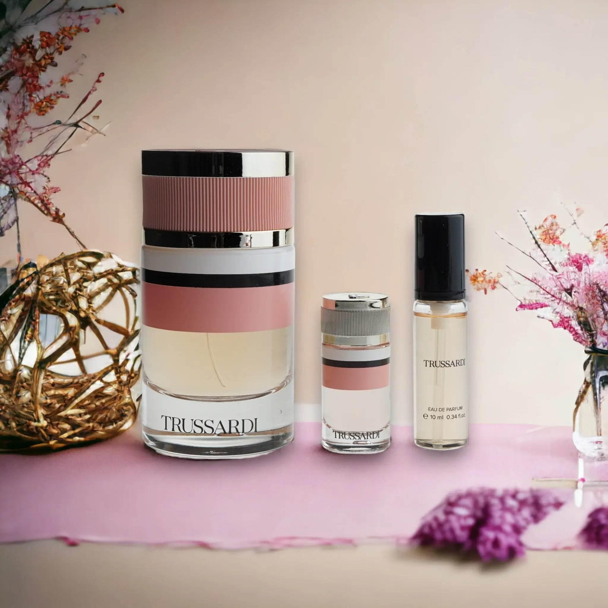 Trussardi Duftset mit drei eleganten Parfumflakons auf rosa Untergrund, Blumen im Hintergrund